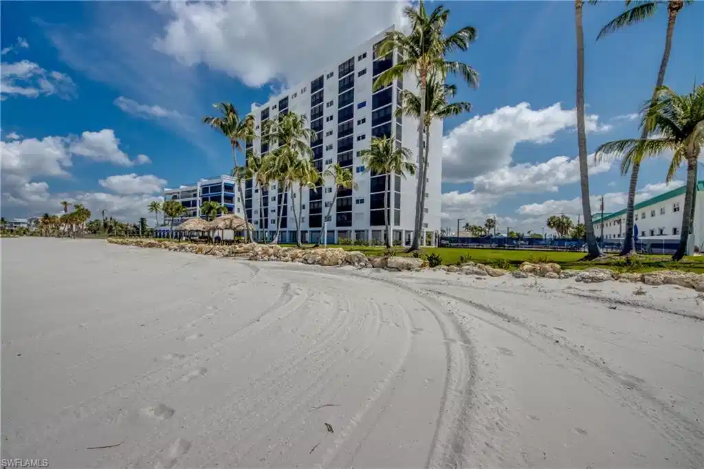 Image 1: Expansive sandy beach with a high-rise building backdrop, featuring numerous palm trees, a thatched-roof structure, and a manicured lawn area
