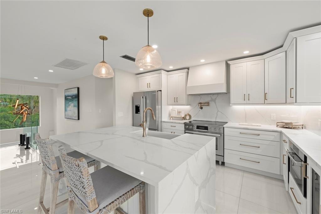 Image 4: Kitchen featuring light stone counters, hanging light fixtures, appliances with stainless steel finishes, an island with sink, and custom range hood