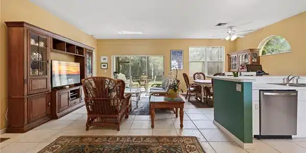 Image 4: Light-toned tile flooring extends throughout this open-concept area featuring a kitchen peninsula with stainless steel appliances, a ceiling fan, and multiple windows providing ambient illumination