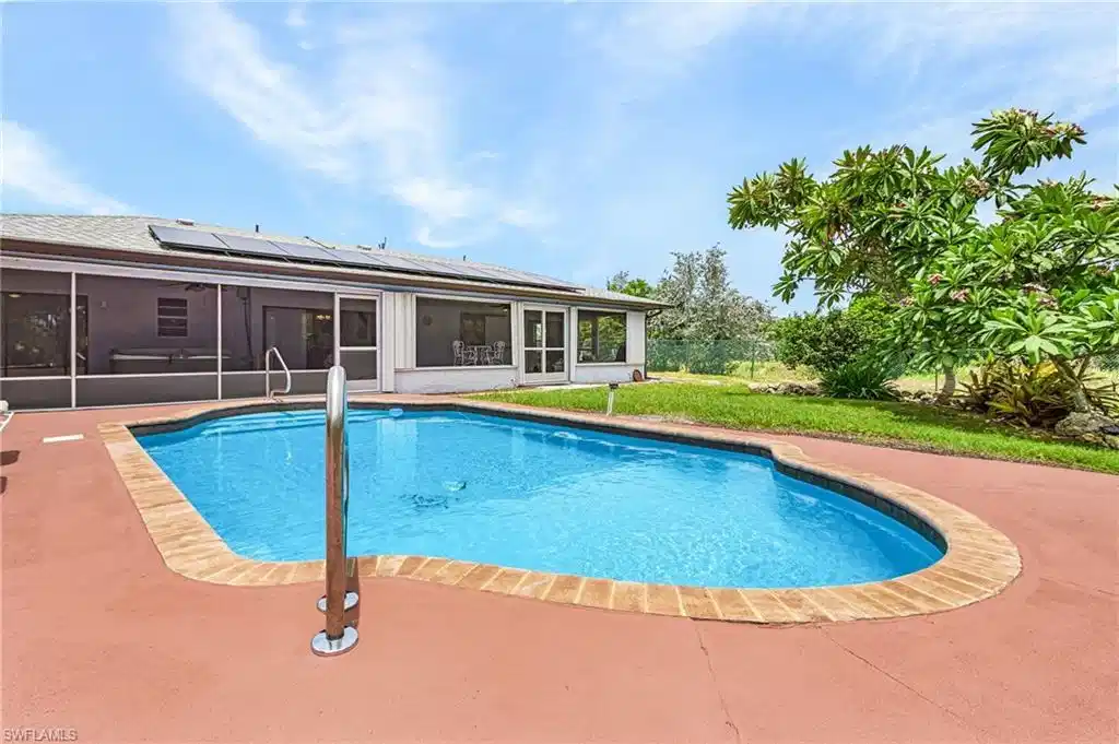 Image 1: Freeform swimming pool with a red-toned concrete deck, brick-patterned coping, and a stainless steel handrail
