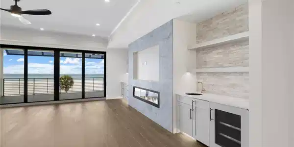 Image 4: Wood-finish flooring throughout, full-length glass doors to the exterior, linear fireplace with a stone-finish surround, built-in wet bar with a beverage cooler, and recessed lighting