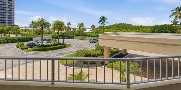 Image 4: Balcony with metal railing overlooking a paved circular drive, palm trees, and distant water