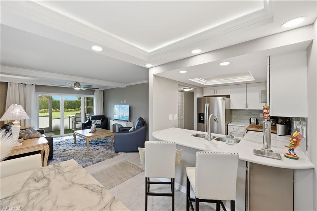 Image 2: Open-concept living area featuring tray ceilings with recessed lighting, sliding glass doors to outdoor space, a kitchen island with seating, stainless steel appliances, and a tile backsplash