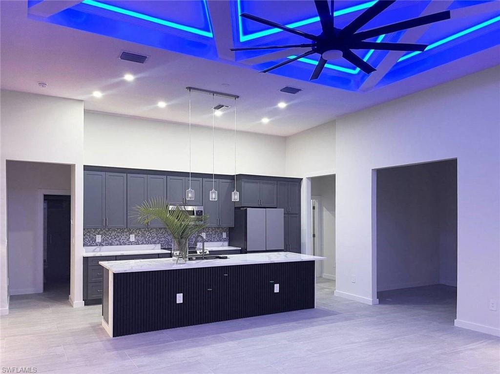 Image 2: Open-concept kitchen featuring an oversized island with a white countertop and pendant lighting, dark gray cabinetry, stainless steel appliances, and a mosaic tile backsplash