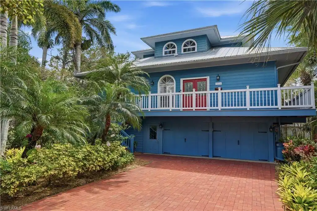 Image 1: Coastal-style home featuring a blue facade with red entry doors, a white railing balcony, and a red brick paver driveway