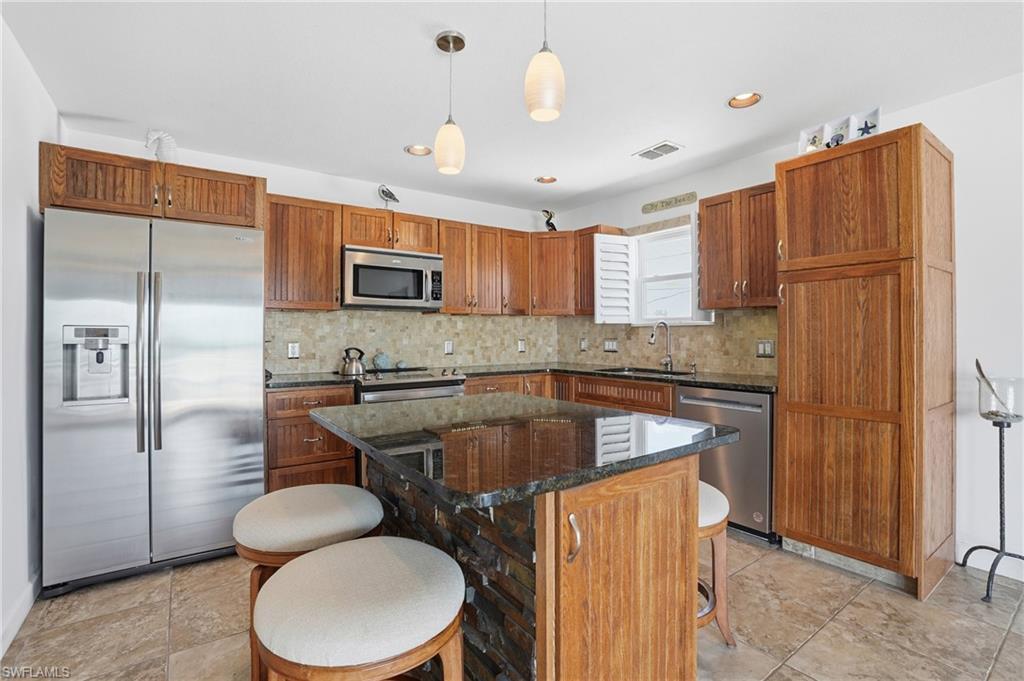 Image 3: Kitchen featuring wood cabinetry, stainless steel appliances, a stone tile backsplash, a dark countertop island with stone detailing, and recessed lighting