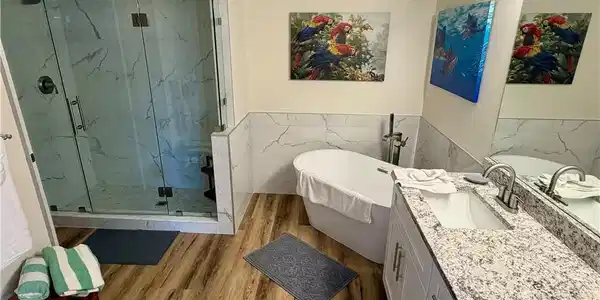 Image 4: Modern bathroom featuring a frameless glass shower enclosure with white marbled tile surround, a freestanding soaking tub, a single vanity with a light-colored speckled countertop, wood-finish flooring, and a large wall-mounted mirror