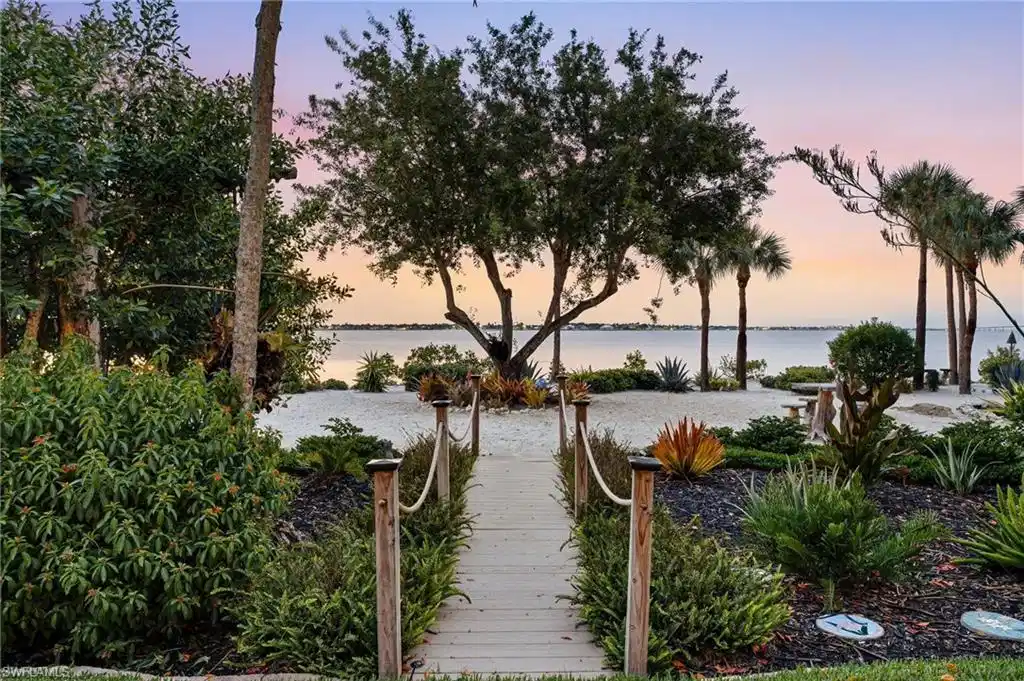 Image 1: A beautifully crafted wooden walkway leads to an exquisitely landscaped private beach, where lush tropical plantings, a sophisticated irrigation system, and striking sculptures create an enchanting waterfront retreat.
