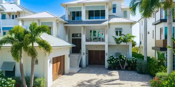 Image 3: Multi-level coastal residence featuring a paver driveway, multiple garage doors with wood-finish panels, a second-story balcony, mature palm trees, and lush landscaping - Virtually Edited Image