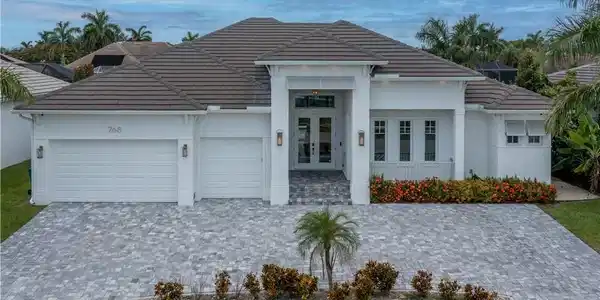 Image 2: Modern residence featuring a striking pitched roof with dark tile detailing, clean white exterior with architectural pillars framing the entrance, triple-car garage configuration, and a paver driveway with manicured landscaping including flowering shrubs and palm trees