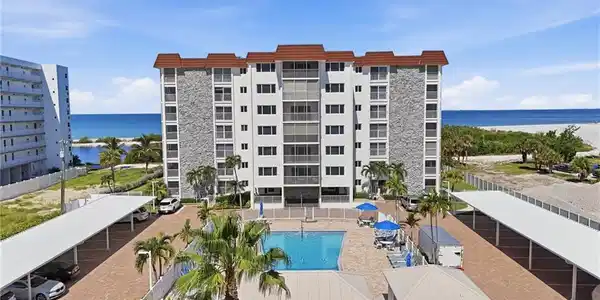 Image 2: Beachfront residential complex featuring a resort-style saltwater pool with surrounding paved deck, mature palm landscaping, and direct oceanfront access - Virtually Edited Image