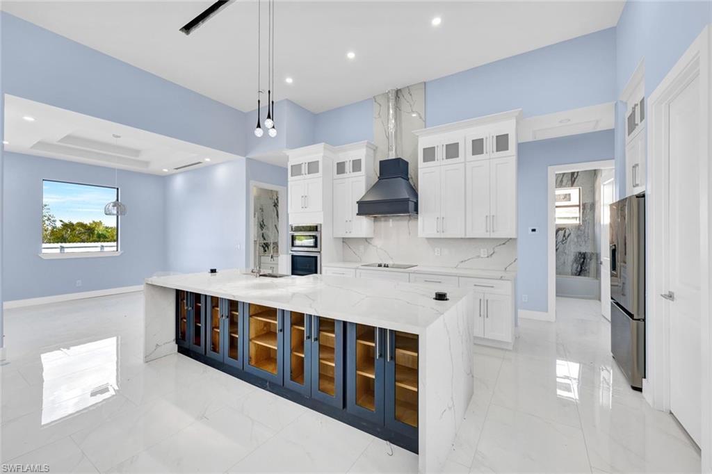 Image 4: Kitchen featuring glass fronted cabinets, white cabinetry, a large island with sink, light stone countertops, and pendant lighting