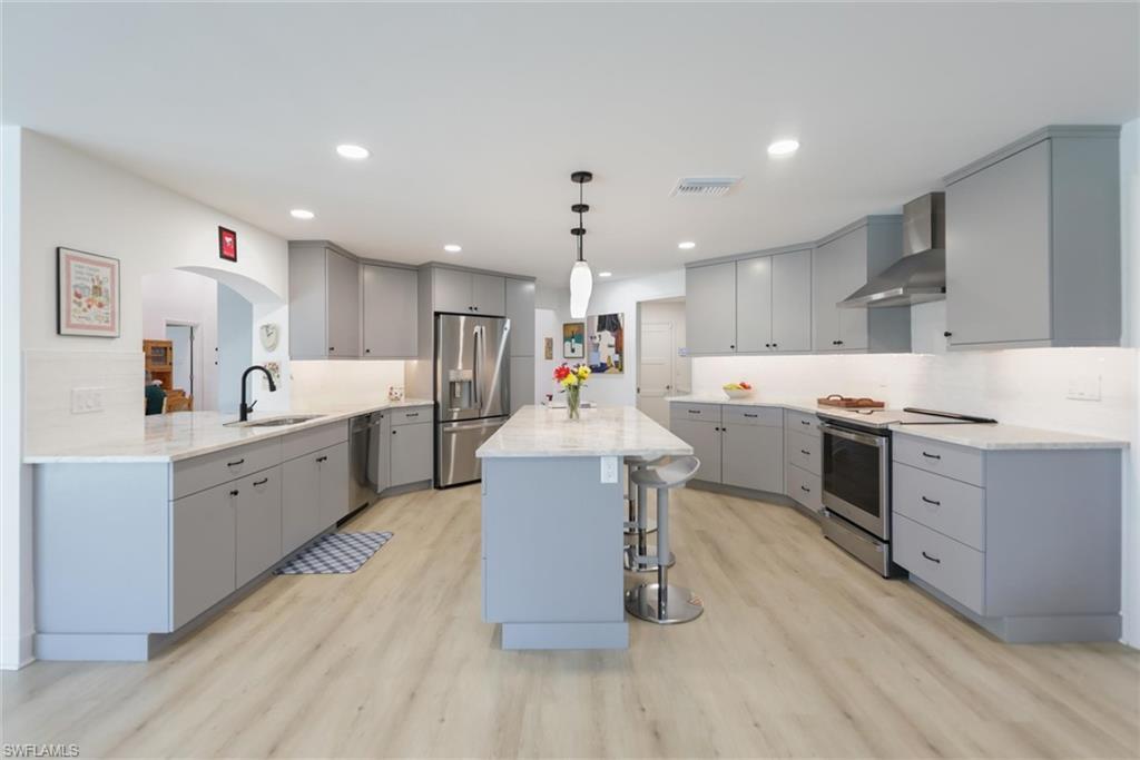 Image 3: Kitchen featuring gray cabinetry, a kitchen breakfast bar, appliances with stainless steel finishes, wall chimney exhaust hood, and hanging light fixtures