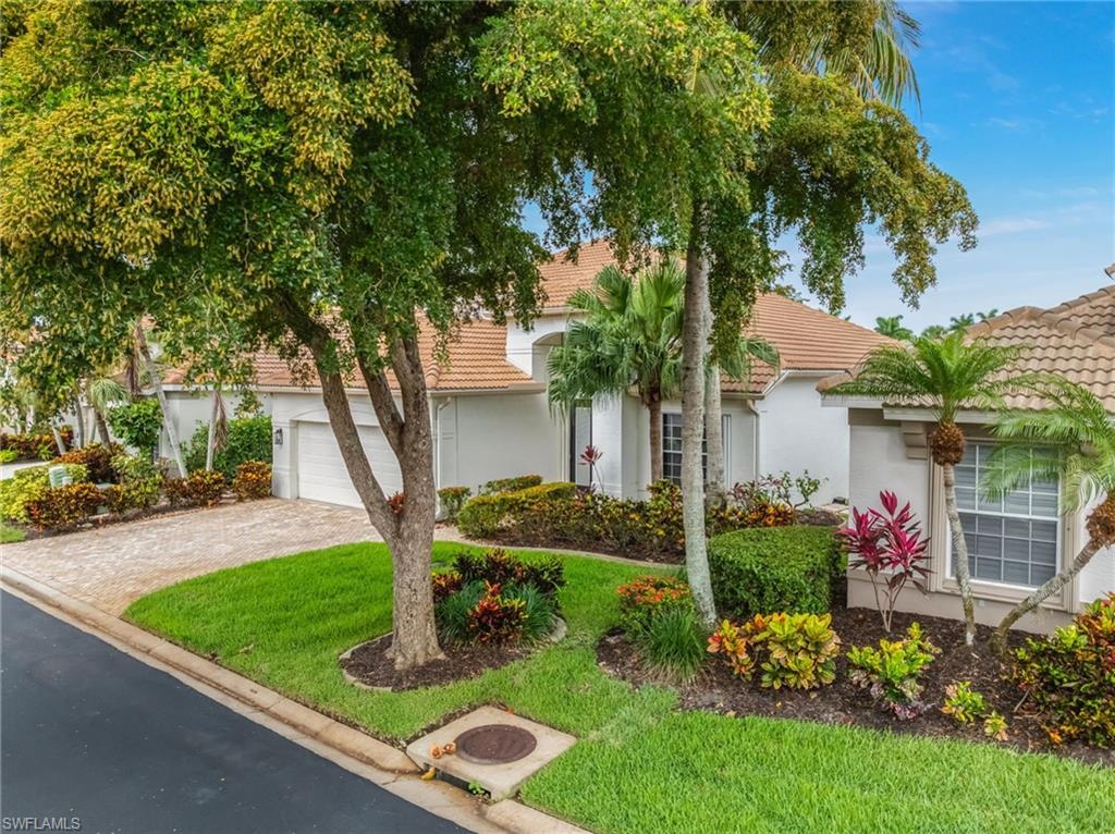 Image 4: Tile roof residence with a paver driveway, an integrated garage, and a lush front yard featuring mature trees, shrubs, and colorful plantings