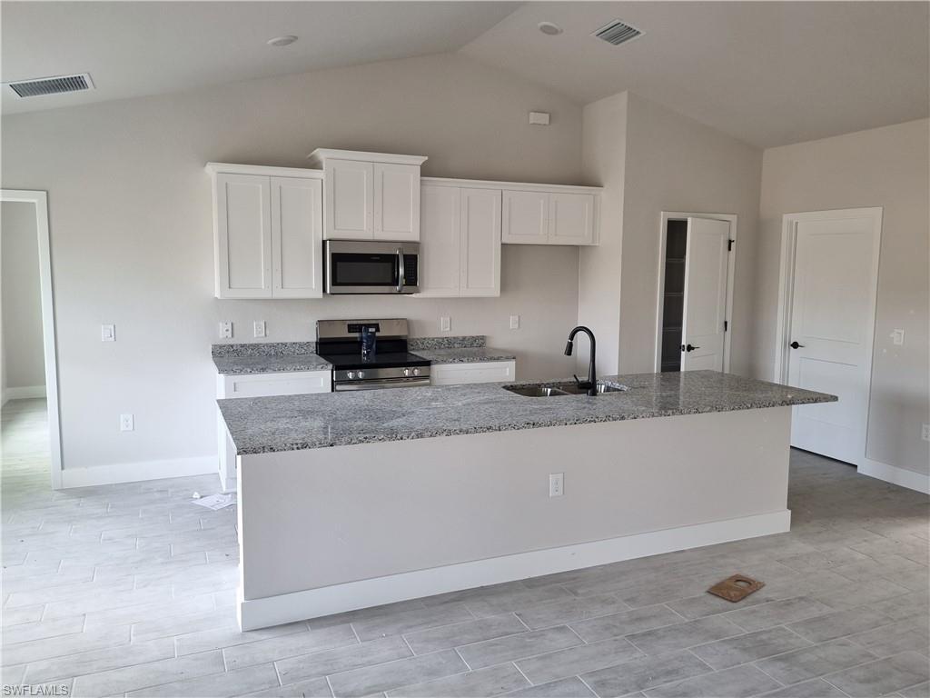 Image 3: Kitchen featuring stainless steel appliances, light stone counters, white cabinetry, an island with sink, and lofted ceiling