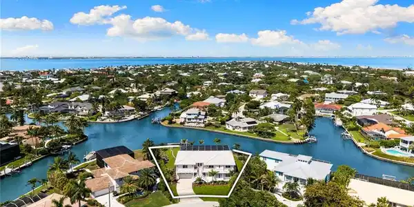 Image 3: Waterfront property featuring a white multi-story exterior, an attached garage, a paved driveway, mature landscaping, and solar panels