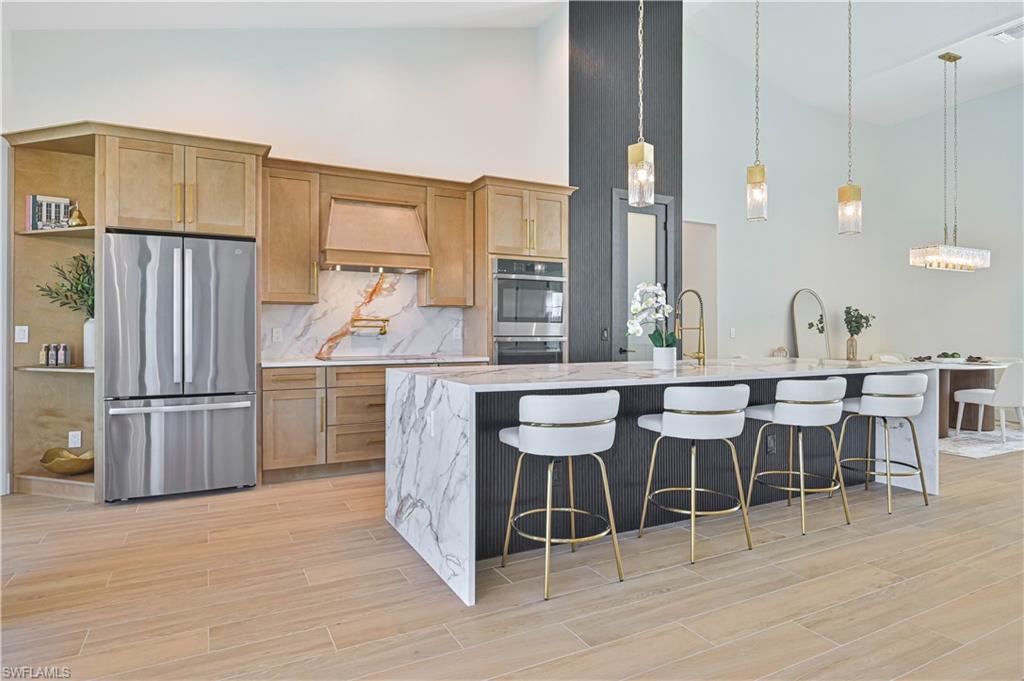 Image 3: Modern kitchen featuring a waterfall island with a light-toned countertop, a built-in stainless steel refrigerator, light wood-finish cabinetry, a matching wood-finish range hood, and plank-style flooring
