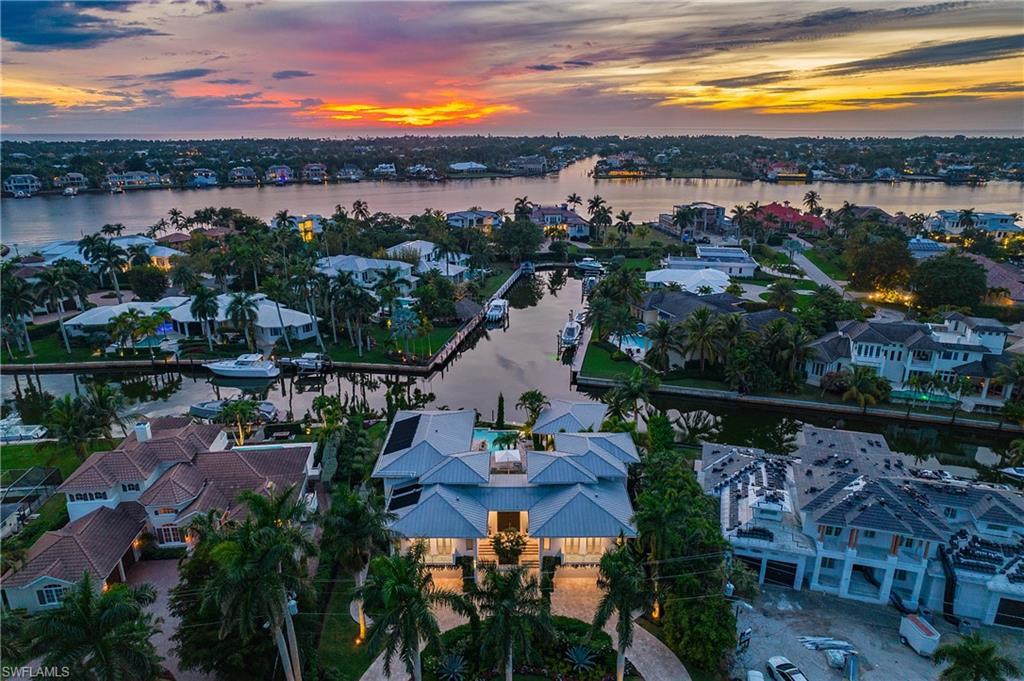 Image 1: Waterfront estate showcasing a striking metal roof, illuminated fa&ccedil;ade, and lush tropical landscaping with mature palms