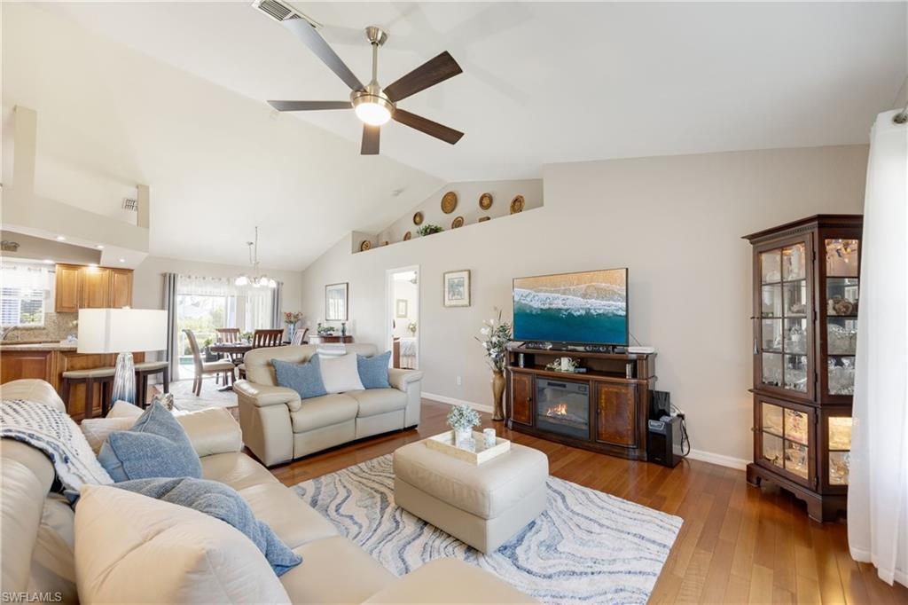 Image 4: Living area with light wood finished floors, a ceiling fan, vaulted ceiling, a glass covered fireplace, and a chandelier