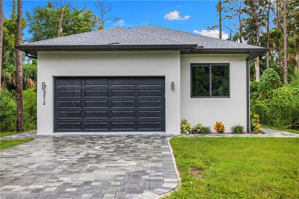 Image 3: Contemporary facade featuring a dark gray paneled garage door, white stucco exterior, black-framed window, and a paver driveway