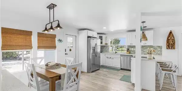 Image 2: Open-concept living space with wood-finish flooring, white cabinetry, stainless steel appliances, a multi-tone tile backsplash, and a dining area with a linear pendant light