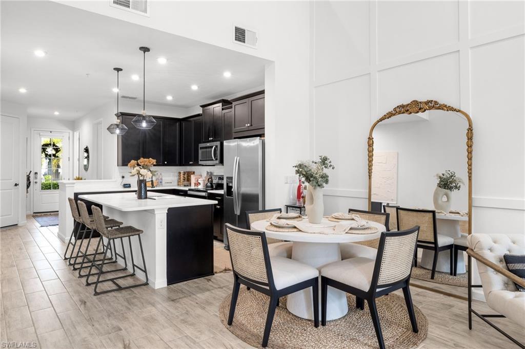 Image 3: Open-concept living space featuring wood-finish flooring, a kitchen with black cabinetry and stainless steel appliances, a kitchen island with seating, a dining area, and a white paneled wall