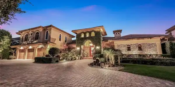 Image 2: Mediterranean-inspired architecture featuring a paver driveway, three-car garage with wood doors, stone facade accents, and lush landscaping with up-lighting