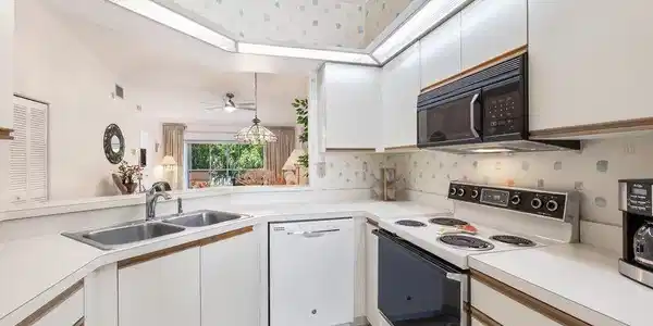 Image 4: Kitchen featuring white cabinetry with wood-finish trim, a double basin stainless steel sink, and white countertops.