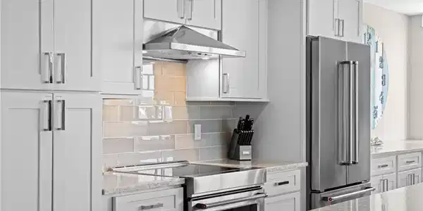 Image 4: Modern kitchen featuring light gray shaker-style cabinetry, stainless steel appliances, a range hood, and a light-toned tile backsplash