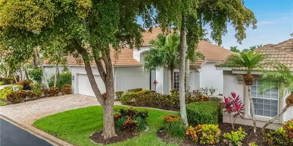 Image 4: Tile roof residence with a paver driveway, an integrated garage, and a lush front yard featuring mature trees, shrubs, and colorful plantings