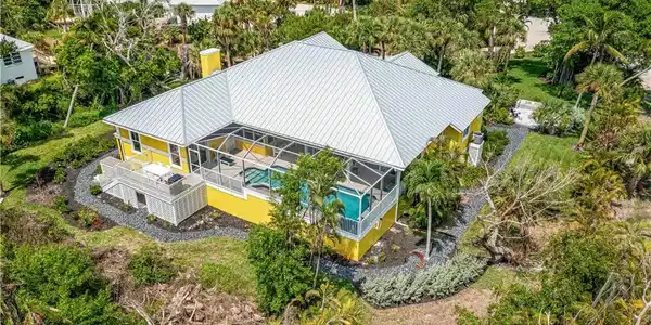 Image 2: Yellow exterior residence featuring a screened-in pool enclosure, a metal roof, an elevated deck, and surrounding landscaping with mature trees and shrubs