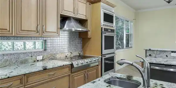 Image 2: Kitchen featuring light wood cabinetry, granite countertops, tile backsplash, built-in stainless steel appliances, and a glass block window accent