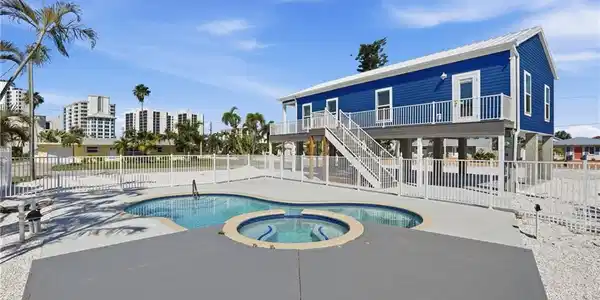 Image 2: Elevated coastal property featuring a blue exterior with white trim, a multi-tiered pool and spa, and a surrounding privacy fence - Virtually Edited Image