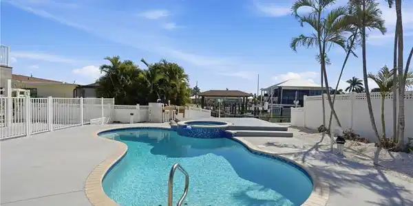 Image 4: Resort-style pool area featuring a freeform swimming pool with integrated spa, surrounding concrete patio, and white privacy fencing - Virtually Edited Image