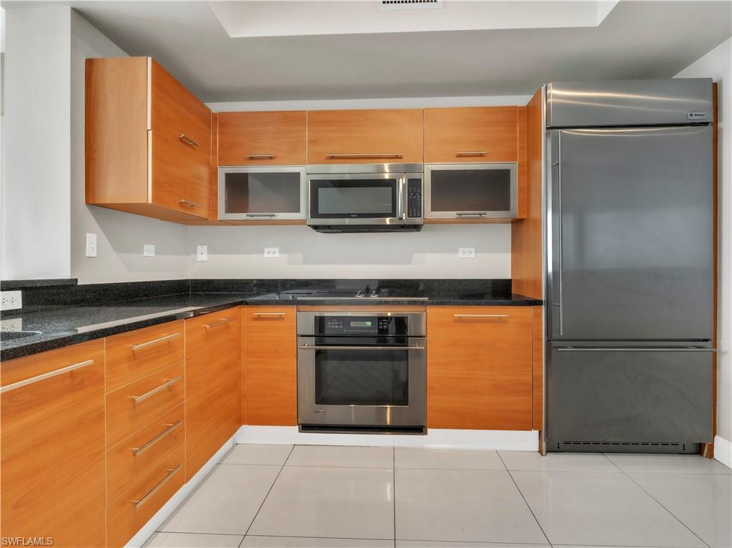 Image 3: Kitchen featuring appliances with stainless steel finishes, light tile patterned flooring, dark stone countertops, and brown cabinets