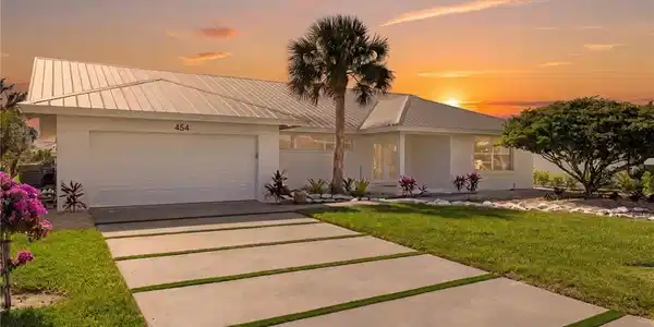 Image 2: The property features a white exterior with a metal roof, a two-car garage, and a concrete driveway with grass strips