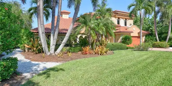 Image 4: Tropical-inspired landscaping frames this Mediterranean-style residence, featuring mature palm trees, lush greenery, and a manicured lawn