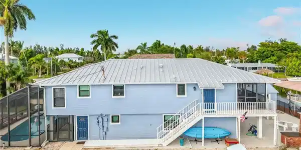 Image 3: Two-story residence with a metal roof, screened-in pool enclosure, exterior staircase, screened-in upper-level porch, and waterfront access with a boat canopy