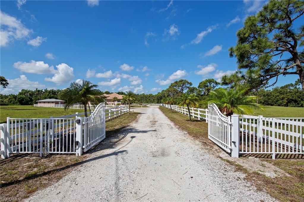 Image 2: Gated entry with white fencing and a gravel driveway, framed by mature trees and expansive green space