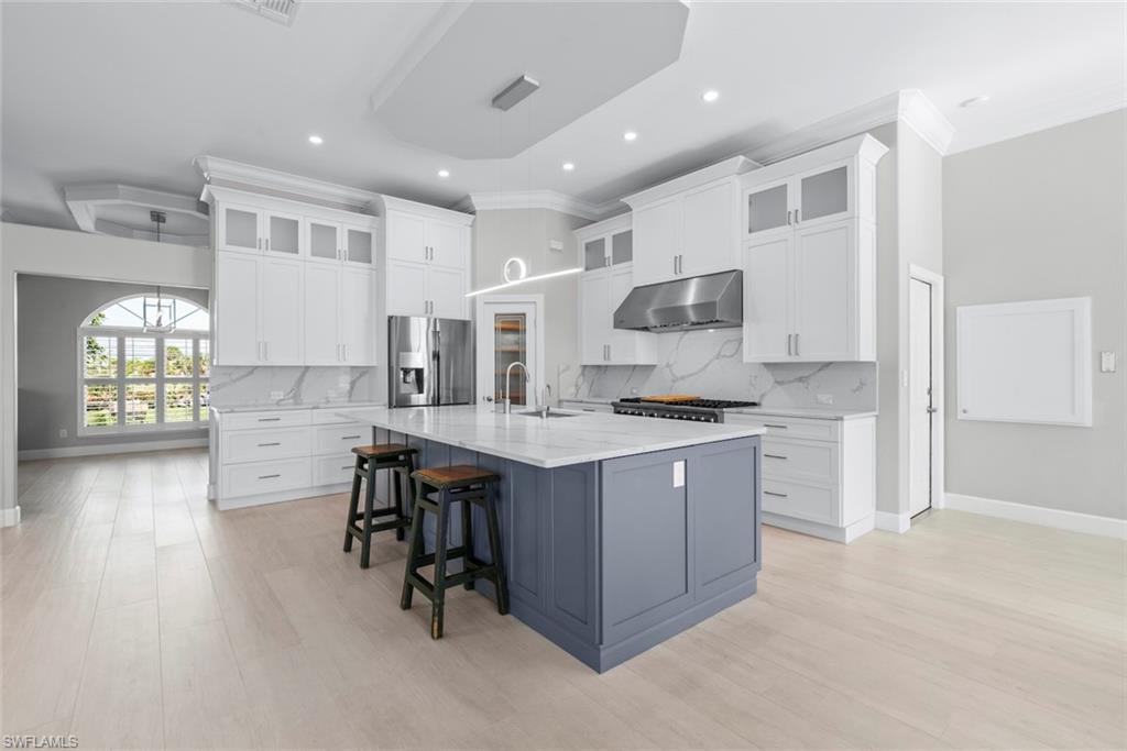 Image 4: Contemporary kitchen featuring a large central island, light wood-finish flooring, white upper and lower cabinetry, stainless steel appliances, and a tray ceiling