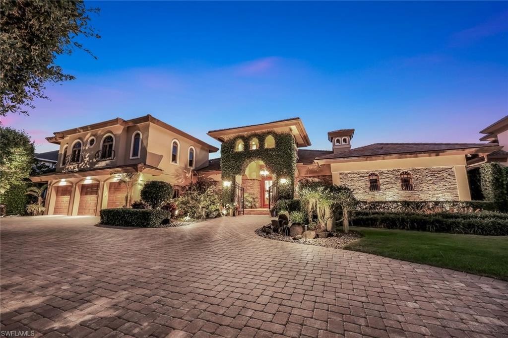 Image 4: Mediterranean-inspired architecture featuring a paver driveway, three-car garage with wood doors, stone facade accents, and lush landscaping with up-lighting