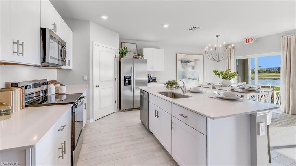 Image 4: Model home. Kitchen featuring an island with sink, stainless steel appliances, white cabinetry, a chandelier, and light stone countertops