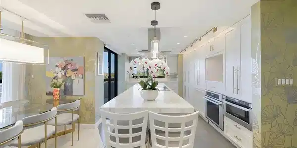 Image 2: Spacious kitchen featuring a large central island with a light-toned countertop, extensive white cabinetry with gold-toned hardware, double wall ovens, and pendant lighting
