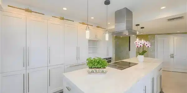Image 4: Gourmet kitchen featuring a substantial island with integrated cooktop, stainless steel range hood, white shaker cabinetry with gold-toned hardware, recessed lighting, and pendant lights