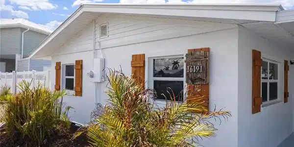 Image 2: Exterior featuring white siding and block construction, wood-finish shutters, white-framed windows, a decorative address plaque, and manicured landscaping