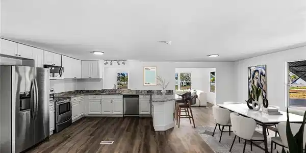 Image 4: Open-concept kitchen and dining area featuring stainless steel appliances, white cabinetry with black countertops, a large island with seating, wood-finish flooring throughout, recessed lighting, and abundant natural light from multiple windows and glass doors