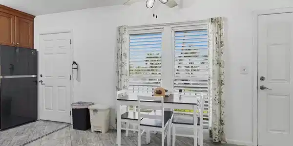 Image 4: Kitchen area featuring light gray tile flooring with a dark accent border, double windows with horizontal blinds, overhead cabinetry, and a ceiling fan with integrated lighting