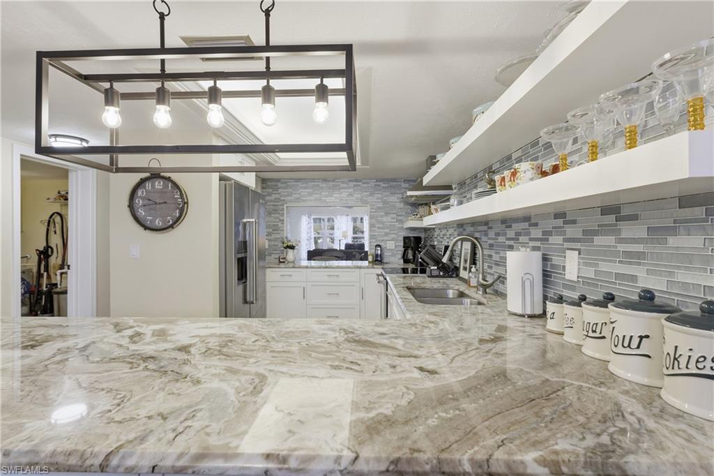 Image 4: Spacious kitchen featuring a large island with a natural stone countertop, a modern rectangular pendant light fixture, a mosaic tile backsplash, white cabinetry, and stainless steel appliances