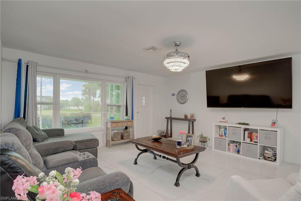Image 3: Spacious living area featuring large windows, white tile flooring, a contemporary ceiling light fixture, a wall-mounted television, and a built-in shelving unit