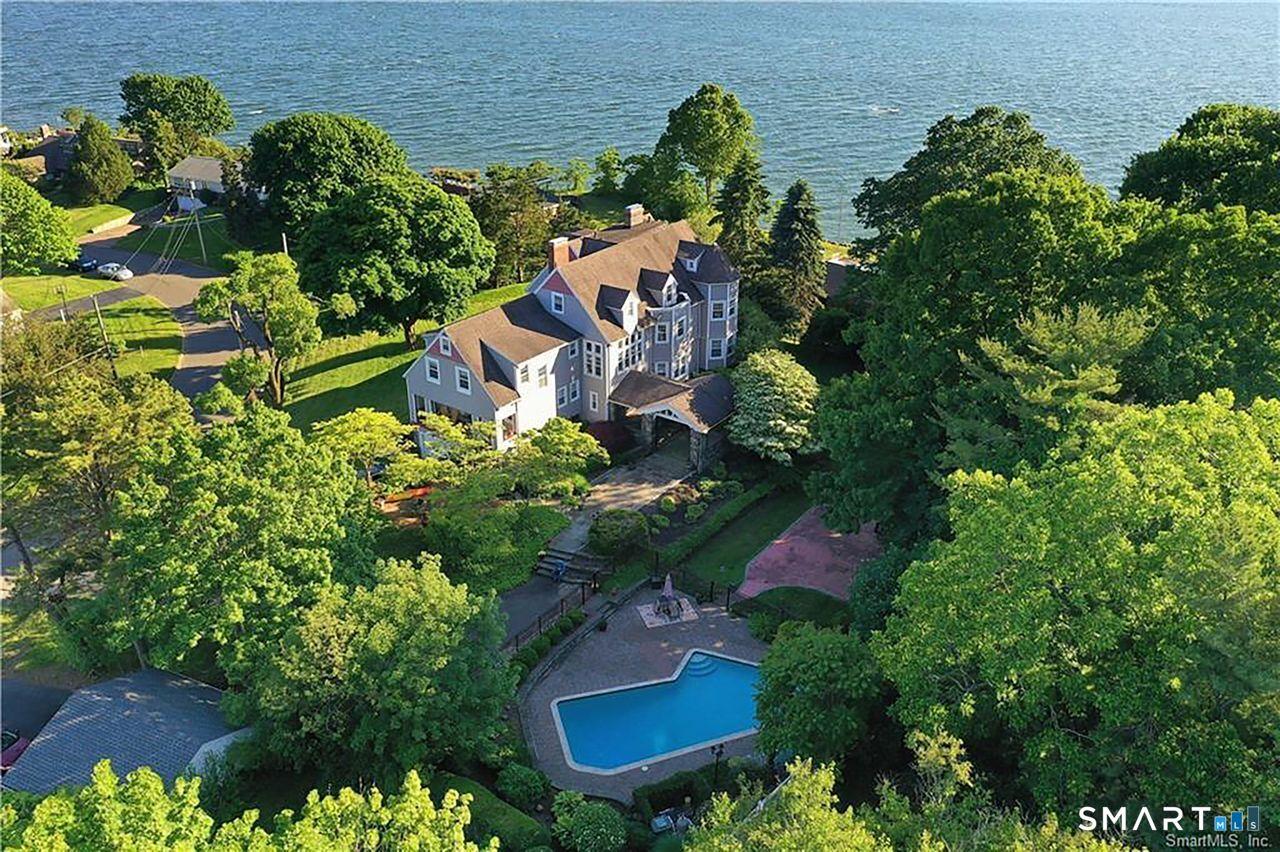 Image 1: A breathtaking aerial view of the estate nestled among mature trees, showcasing its elevated setting, heated pool, and expansive grounds, all framed by sweeping vistas of Long Island Sound in three directions.